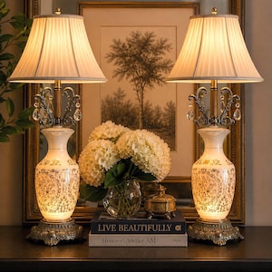 May include: Two matching cream-colored table lamps with pleated shades and ornate bases. The lamps have a floral design and crystal accents. A vase of white flowers and books with the text "LIVE BEAUTIFULLY" are between the lamps.