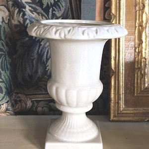 May include: A white, antique-style urn with a decorative rim and base. The urn has a textured surface and a classic, symmetrical design. It stands on a square base and is suitable for floral arrangements or decorative display.