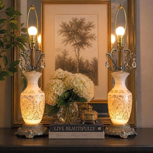 May include: Two matching table lamps with ornate bases and cream-colored, patterned bodies. Each lamp has a gold-toned neck, a light bulb, and decorative crystal accents. The lamps are on a dark surface, with a framed picture and flowers in the background.