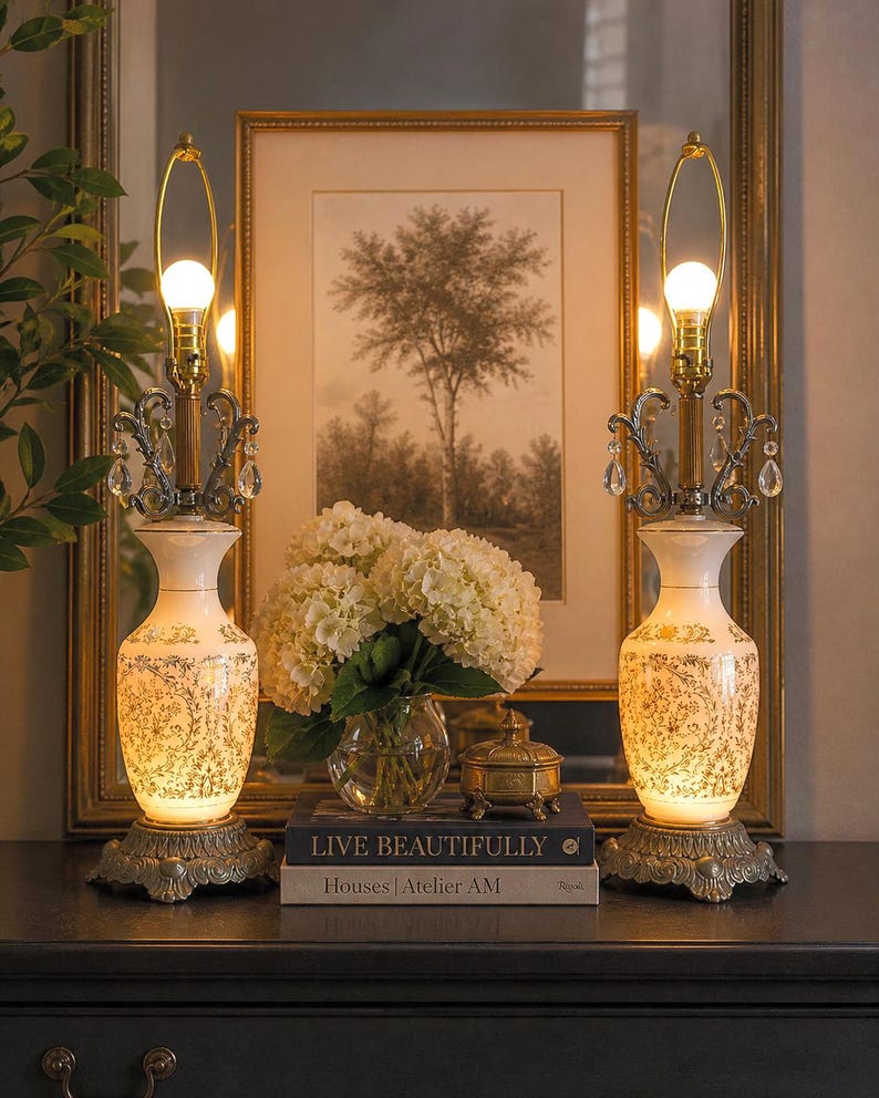 May include: Two matching table lamps with ornate bases and cream-colored, patterned bodies. Each lamp has a gold-toned neck, a light bulb, and decorative crystal accents. The lamps are on a dark surface, with a framed picture and flowers in the background.