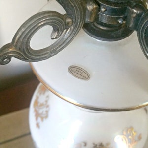 May include: Close-up of a white and gold decorative lamp. The lamp has a white glass shade and a white base with gold floral designs. The metal accents are silver and gold. The label on the lamp reads "BASE MADE IN FRANCE."