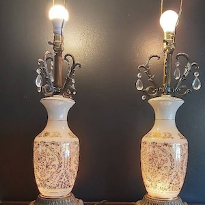 May include: Pair of vintage table lamps. Each lamp features a white vase-shaped base with gold floral patterns. The lamps have ornate metal accents, crystal embellishments, and a gold-toned lamp shade holder. The lamps are lit with warm light.