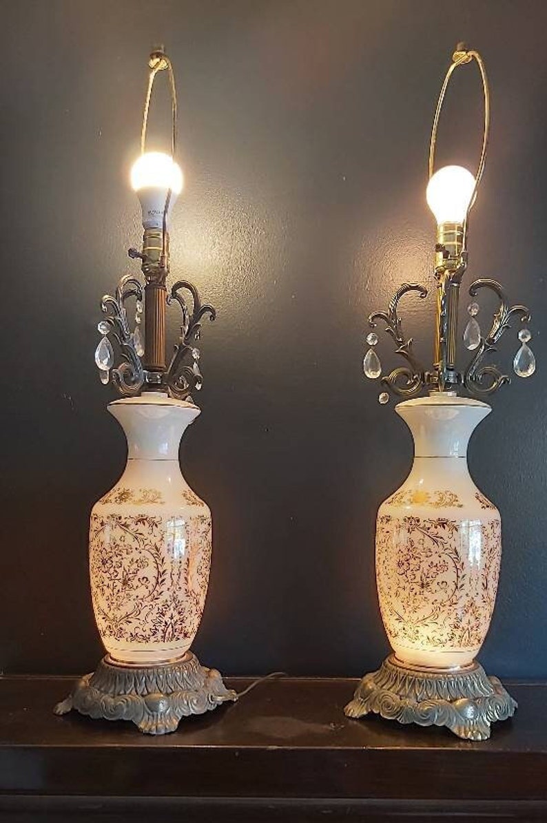 May include: Pair of vintage table lamps. Each lamp features a white vase-shaped base with gold floral patterns. The lamps have ornate metal accents, crystal embellishments, and a gold-toned lamp shade holder. The lamps are lit with warm light.