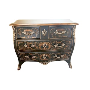 May include: A black antique chest of drawers with four drawers and ornate gold-colored accents. The top of the chest is white, and the edges are trimmed in gold. The drawers have gold-colored handles and decorative floral designs.
