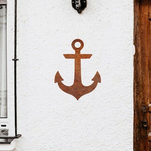 Könnte beinhalten: Eine weiße Außenwand mit einer braunen Holztür und einem Fenster mit schwarzen Eisenstäben. Ein rostiger brauner Anker ist an der Wand über der Tür angebracht. Die Wand hat unten einen dekorativen Fliesenrand.