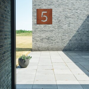 May include: A view through a window of a brick building with a rusty metal number "5" on the wall. The building is surrounded by a paved patio.