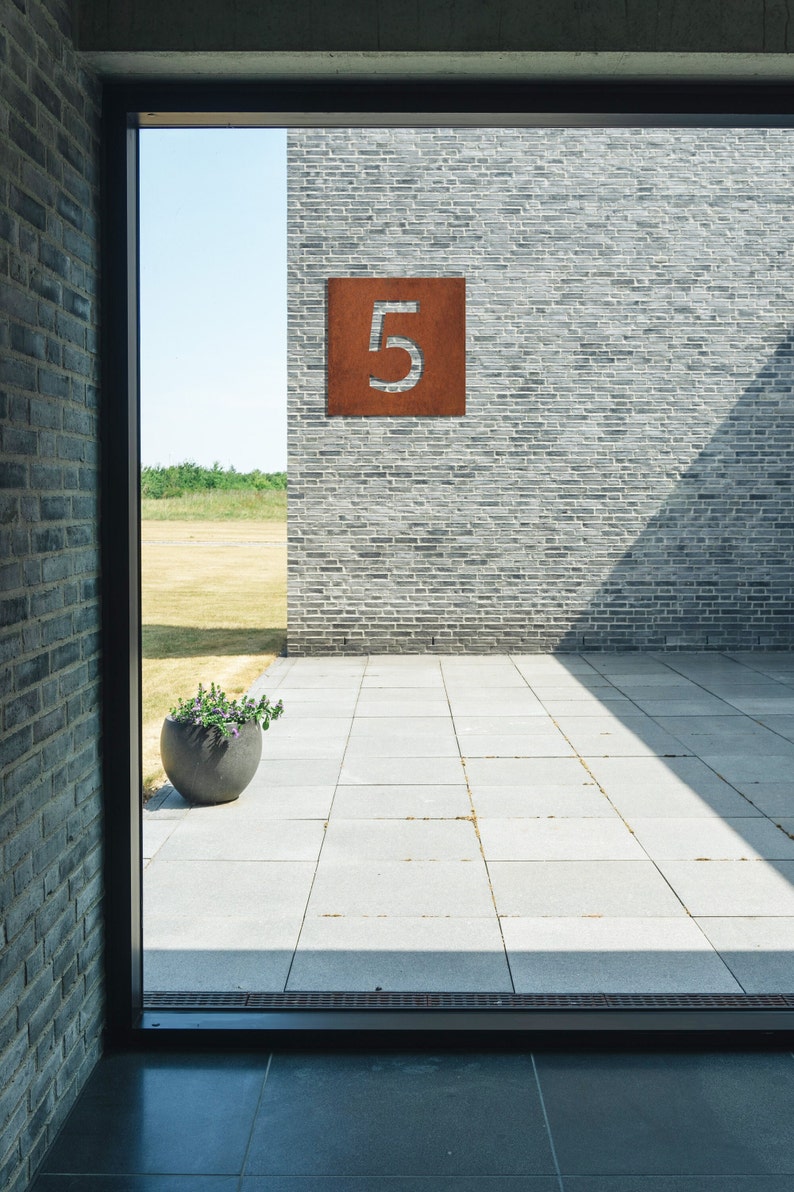 May include: A view through a window of a brick building with a rusty metal number "5" on the wall. The building is surrounded by a paved patio.