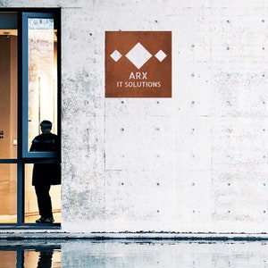 May include: A brown rectangular sign with a white geometric design and the text "ARX IT SOLUTIONS" on a white concrete wall.