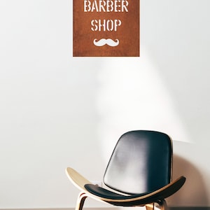 May include: A brown sign with white lettering that reads "BARBER SHOP" and a white moustache underneath. A black leather chair with wooden legs sits on a wooden floor.