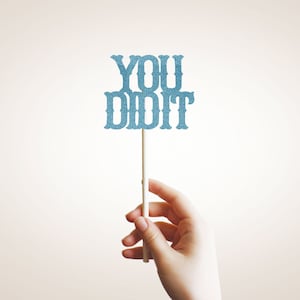 May include: A hand holds a blue glitter sign that says "YOU DID IT" on a stick.