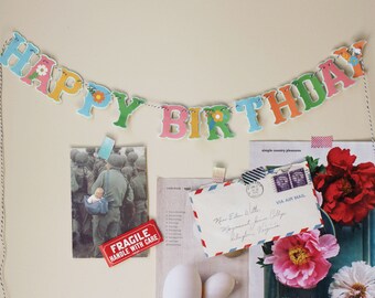 May include: A colorful Happy Birthday banner with a string of letters and flowers. The banner is hanging on a wall with other decorations, including a postcard, a letter, and a photo.