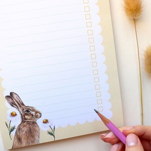 May include: A white notepad with a scalloped edge and a brown rabbit illustration. The notepad has lined paper and a checklist on the right side. A pink pencil is being used to check off items on the list.