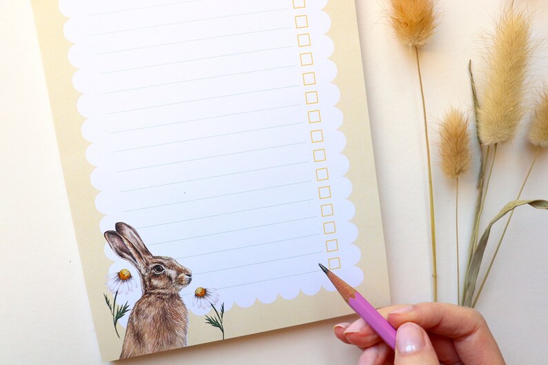 May include: A white notepad with a scalloped edge and a brown rabbit illustration. The notepad has lined paper and a checklist on the right side. A pink pencil is being used to check off items on the list.