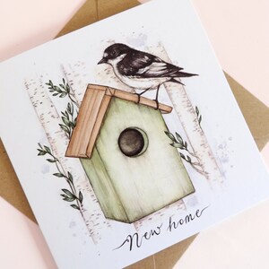 May include: A watercolor illustration of a bird perched on a green birdhouse with a brown roof. The birdhouse is surrounded by birch trees and green leaves. The text "New home" is written in cursive at the bottom of the card.