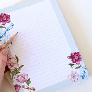 May include: A blue lined notepad with a floral design. The design features pink and white flowers with blue ribbons on a light blue background.