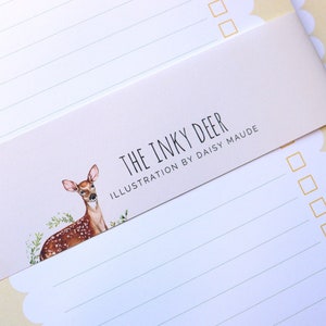 May include: A set of two white lined notepads with a scalloped edge. The notepads have a yellow background and a sticker with a watercolor illustration of a deer and the text "The Inky Deer Illustration by Daisy Maude".
