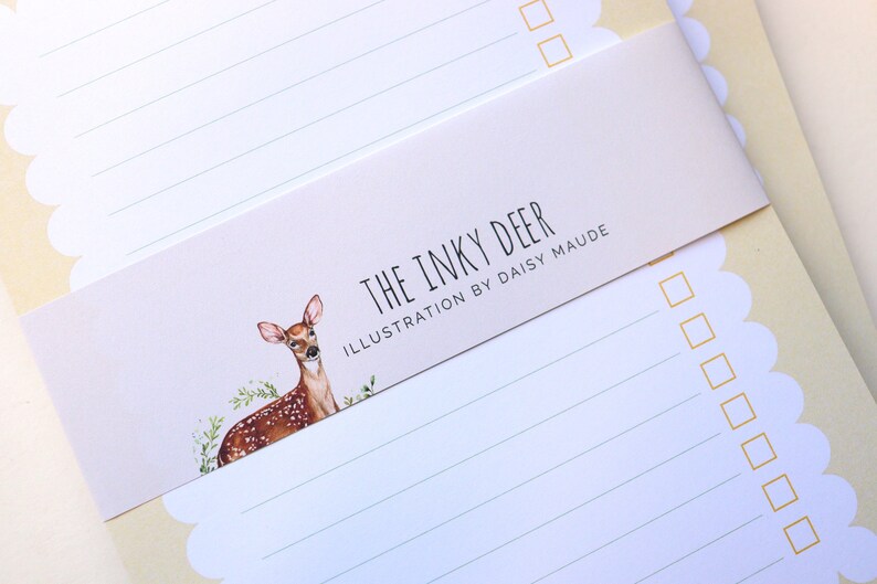 May include: A set of two white lined notepads with a scalloped edge. The notepads have a yellow background and a sticker with a watercolor illustration of a deer and the text "The Inky Deer Illustration by Daisy Maude".