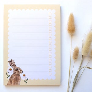 May include: A beige notepad with a scalloped edge and a white interior. The notepad has a line design and a checklist. The notepad features a watercolor illustration of a brown rabbit with white flowers.