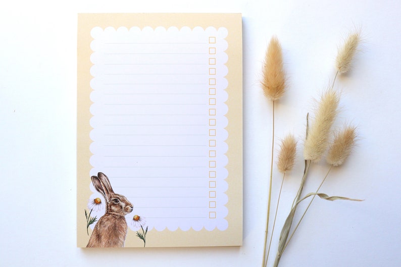 May include: A beige notepad with a scalloped edge and a white interior. The notepad has a line design and a checklist. The notepad features a watercolor illustration of a brown rabbit with white flowers.