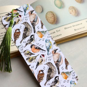 May include: A bird-themed bookmark with a green tassel, resting on an open book. The bookmark showcases detailed illustrations of different bird species on a white backdrop. A delightful accessory for any book enthusiast.