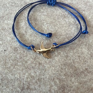 Puede incluir: Un brazalete de cuerda azul con un dije de pájaro dorado. El brazalete está atado con un nudo y tiene un diseño simple.