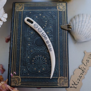 May include: A white bone needle with black circles and a decorative design. The needle is lying on a dark blue book with gold accents. The book is titled "Wizard's Cave".
