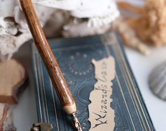May include: A wooden dip pen with a brown and cream colored barrel and a silver nib. The pen is resting on a blue and gold journal with a brown paper tag that says "Wizards Cards" in black calligraphy.
