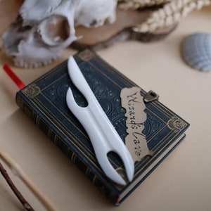 Pode incluir: Um marcador de página branco em forma de espada estilizada com uma alça decorativa, descansando sobre um livro azul escuro com detalhes dourados. O livro tem um marcador de página de papel castanho com o texto "Wizard's Lane" escrito nele.