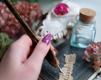 May include: A hand holding a wooden dip pen with a metal nib, writing the words "Wizard's Cave" on a brown paper tag. The tag is placed on a dark blue book with gold accents. A small glass bottle with a cork stopper and a white animal skull with a pink flower inside are in the background.