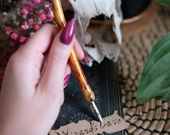 May include: A hand holding a brown and gold calligraphy pen with a silver nib, writing on a brown paper tag with the words "Wizard's Cave" in black ink. The tag is on top of a dark green book with gold accents.
