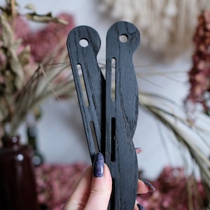 May include: Two black wooden knitting needles with a curved shape and a hole at the top. The needles are held in a hand.