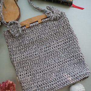 Puede incluir: Un bolso de crochet gris con un asa de madera y un diseño floral. El bolso está sobre una superficie gris claro con una concha y otros elementos decorativos.