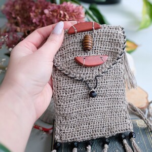Puede incluir: Bolso de teléfono de crochet beige hecho a mano. El bolso presenta detalles de madera y piedra, incluyendo una cuenta de madera central y dos piezas de piedra marrón rojizo. Cuentas negras y borlas adornan la parte inferior, con una correa larga.