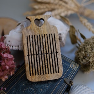 May include: A wooden comb with a heart-shaped cutout at the top. The comb has a natural wood finish and is made of a light brown wood. The comb is sitting on a blue book with a gold clasp.