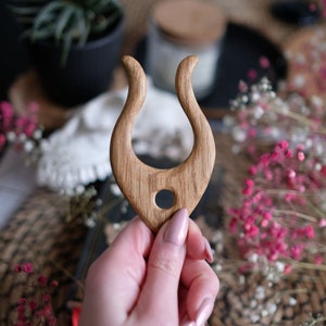 Op de afbeelding: Een houten haarvork met een uniek, gebogen ontwerp. De vork is gemaakt van lichtgekleurd hout en heeft een centraal gat. De algemene vorm doet denken aan een gestileerde U of een paar hoorns. De achtergrond omvat bloemen en een geweven mat.