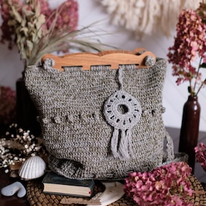 Puede incluir: Una bolsa de mano de crochet gris con un asa de madera y un amuleto de atrapasueños de crochet gris. La bolsa está sobre una estera tejida con flores secas y un libro.
