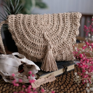 Puede incluir: Una bolsa de yute tejida a crochet con un borla, sentada sobre una estera tejida con un libro y una pequeña calavera de animal. El libro tiene una etiqueta que dice "Wizard's Cave".