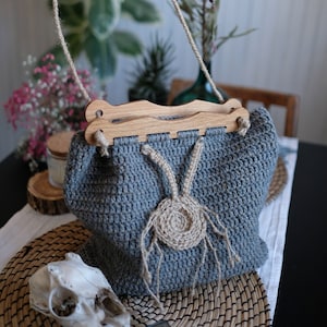 Puede incluir: Una bolsa de crochet gris con un asa de madera y un detalle de crochet marrón. La bolsa está sentada sobre un mantel tejido.