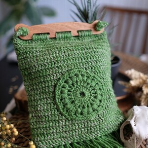 Puede incluir: Bolso de crochet verde con asa de madera y flecos. El bolso tiene un diseño circular en el centro.