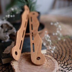 Può includere: Due supporti per cinturino di chitarra in legno, uno sopra l'altro, con finitura in legno naturale. I supporti sono appoggiati su un ceppo d'albero.