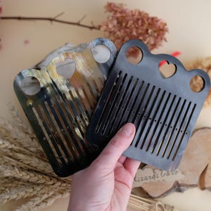 Puede incluir: Dos peines hechos de cuerno, uno negro y otro con un patrón de cuerno natural. Ambos peines tienen un corte en forma de corazón y una fila de dientes.