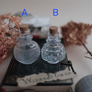 Könnte beinhalten: Zwei kleine, klare Glasflaschen mit Korken. Flasche A hat ein Rautenmuster, während Flasche B ein geometrisches Design hat. Sie stehen auf einem dunklen Buch mit dem Text "Wizard's Cave" auf einem Etikett. Getrocknete Blumen und ein Schlüssel sind ebenfalls vorhanden.