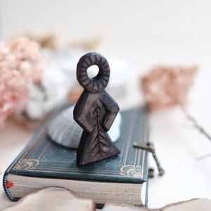 Puede incluir: Una figura de madera tallada de color marrón oscuro con cabeza circular y cuerpo triangular, sobre un libro antiguo. La figura tiene un diseño en forma de hoja en la parte posterior. El libro es azul oscuro con detalles dorados.