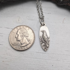 May include: A silver-colored pendant necklace featuring a detailed raven design perched on a branch. The pendant is oval-shaped and hangs from a delicate chain. A quarter coin is next to the pendant for size reference.