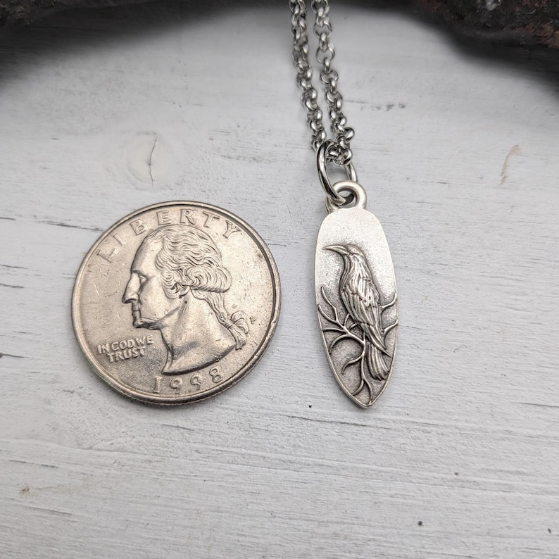May include: A silver-colored pendant necklace featuring a detailed raven design perched on a branch. The pendant is oval-shaped and hangs from a delicate chain. A quarter coin is next to the pendant for size reference.