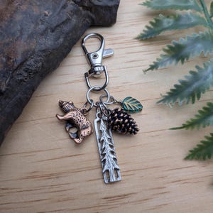 May include: A silver-tone keychain with a lobster clasp, featuring charms of a bear, a pine tree, a pine cone, and a green leaf. The bear charm is bronze-colored. The pine cone is brown. The leaf charm has green and gold accents.