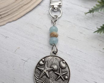May include: A silver-colored keychain with a round pendant featuring starfish and seashells. The keychain has a lobster clasp and is adorned with blue and brown beads. The design evokes a coastal theme.