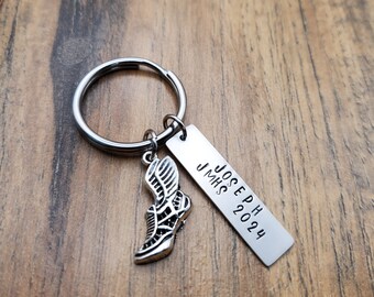 May include: A silver-colored keychain with a running shoe charm and a rectangular tag. The tag is engraved with the name "JOSEPH", "JMHS", and the year "2024". The keychain is on a wooden surface.