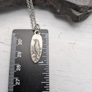 May include: Silver-toned pendant necklace featuring a detailed bird design. The pendant is oval-shaped and hangs from a delicate chain. The pendant is approximately 1 inch long, as measured by the ruler in the image. The bird is perched on a branch.