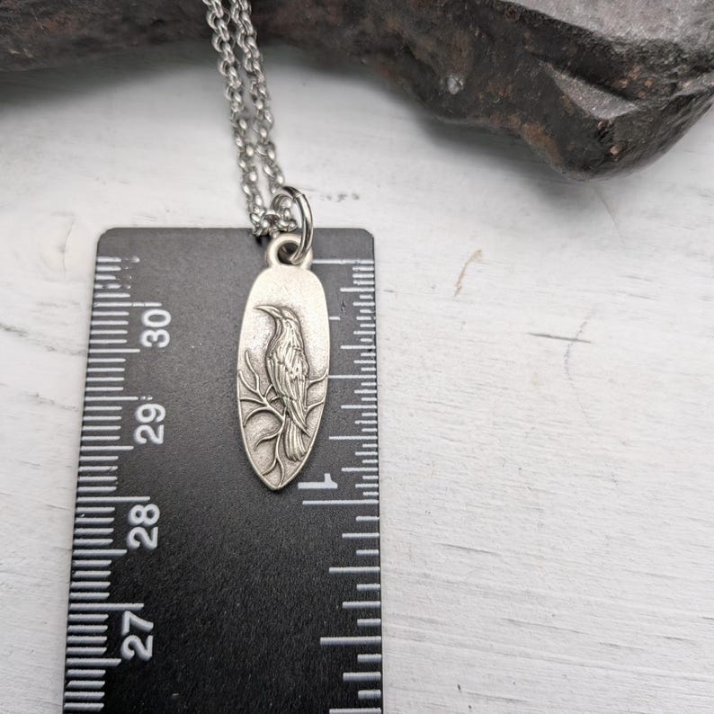 May include: Silver-toned pendant necklace featuring a detailed bird design. The pendant is oval-shaped and hangs from a delicate chain. The pendant is approximately 1 inch long, as measured by the ruler in the image. The bird is perched on a branch.